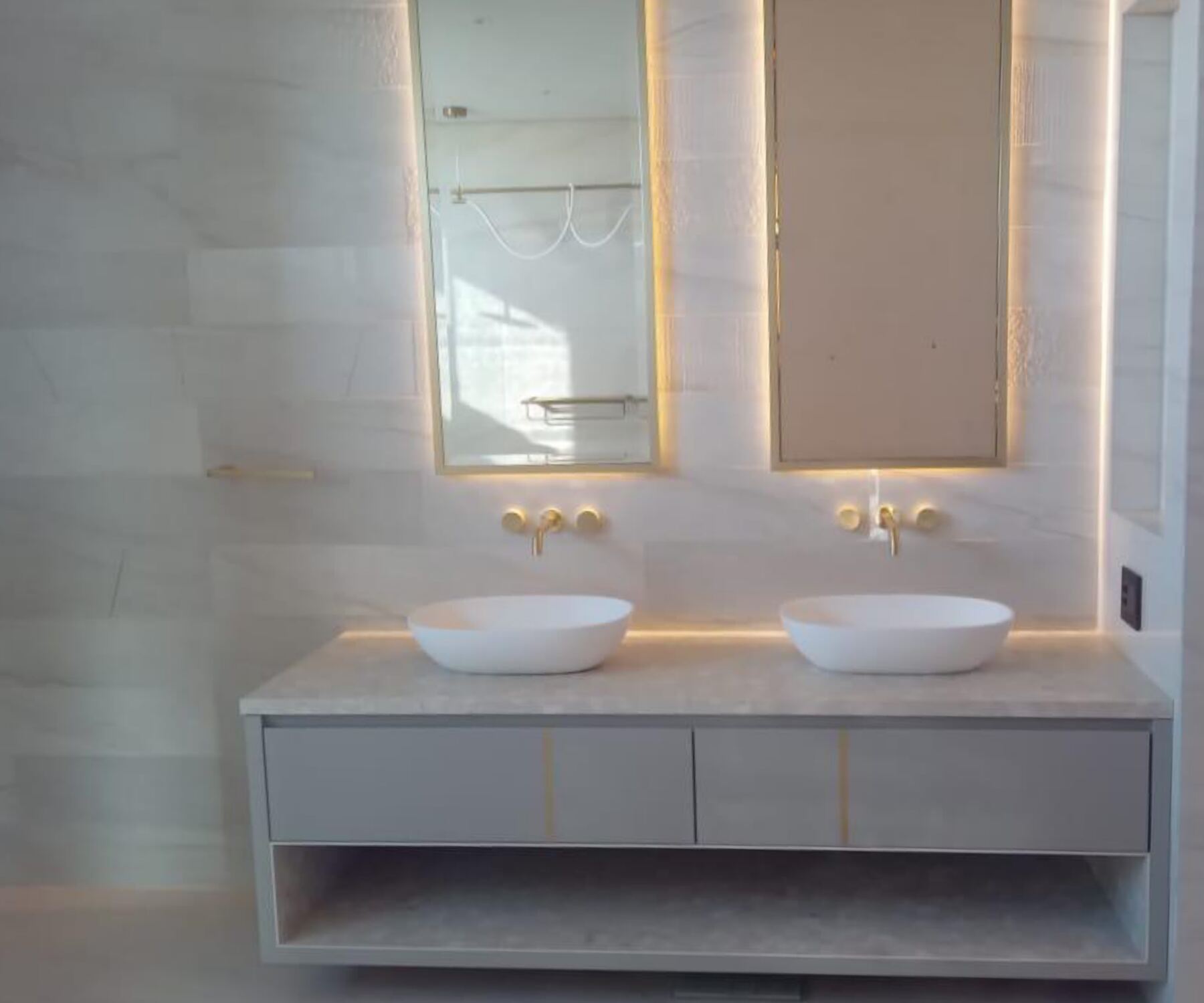 Streamlined contemporary Pandors Designs vanity in soft grey cabinetry with backlit oval mirrors and brass fittings