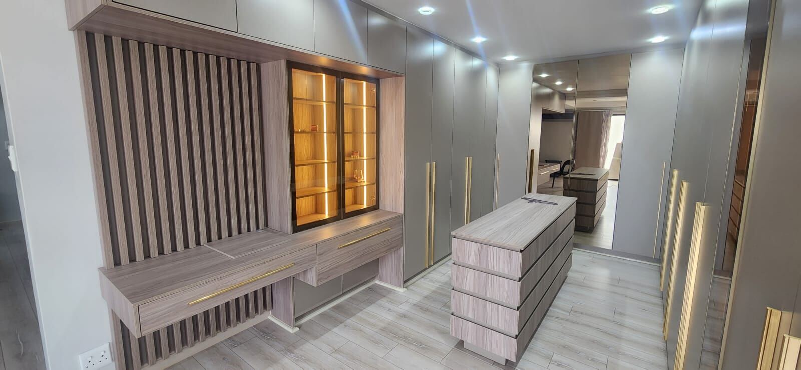 Pandors Designs dressing area with a backlit glass display cabinet, slatted wood panelling and a freestanding drawer island